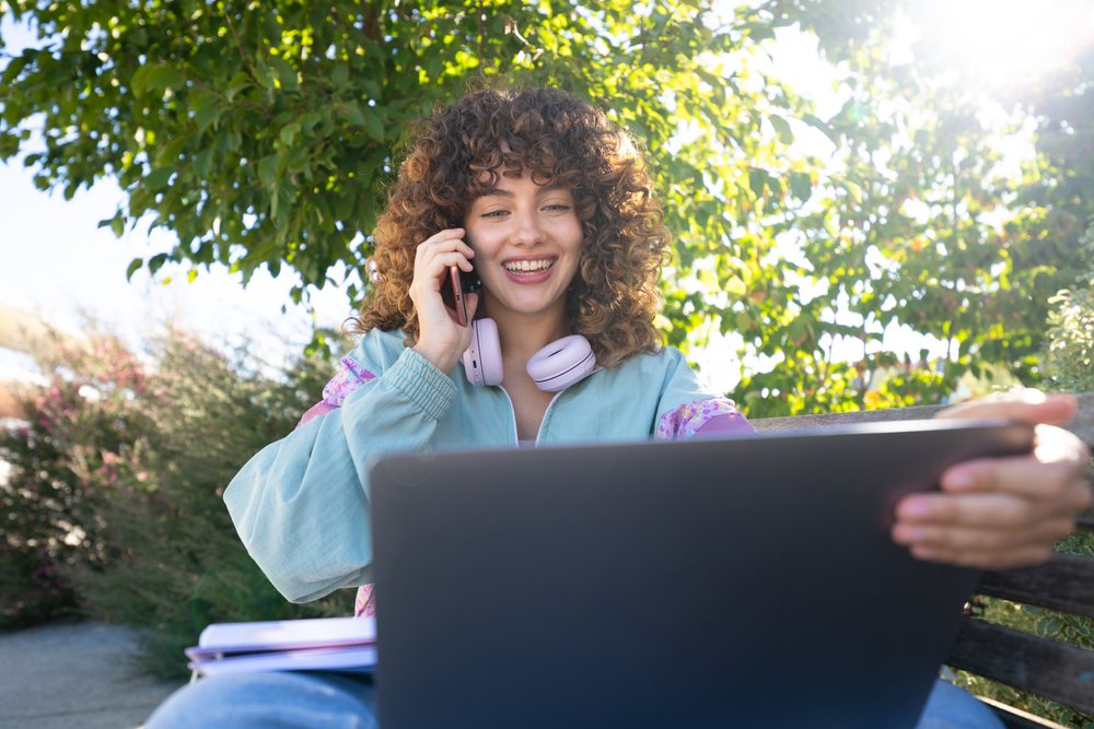 Woman with curly hair smiling while talking on phone and using laptop outdoors on a bench, Spain.