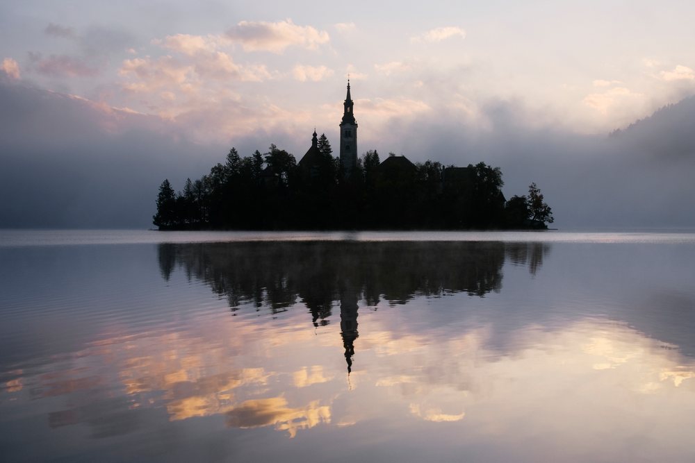 Church of Assumption silhouetted on a foggy lake island with reflection, Lake Bled, Slovenia.