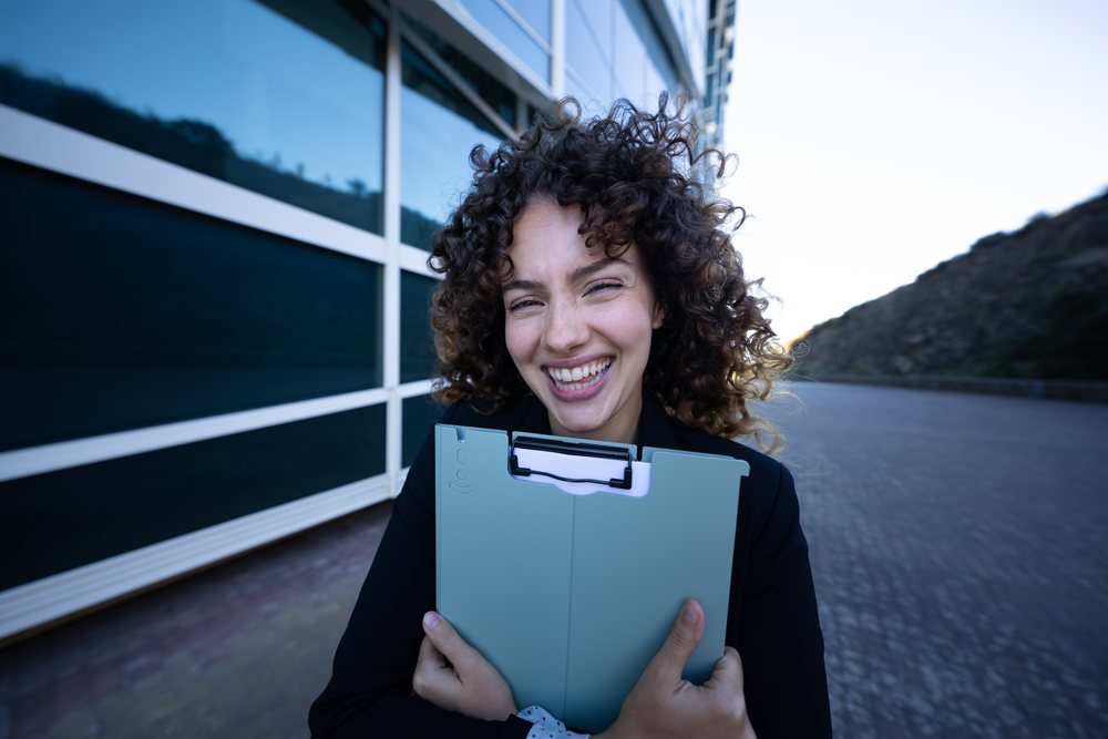 Businesswoman holding a folder and smiling outdoors near a modern building, business trip, Spain, Santiago De Compostela.