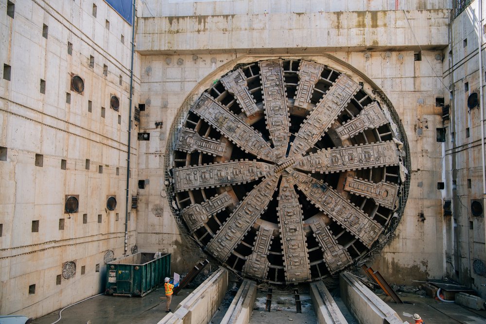 A large tunnel boring machine at a construction site in the USA with a worker in a safety vest inspecting blueprints nearby, large tunnel boring machine, tunnel, drilling.