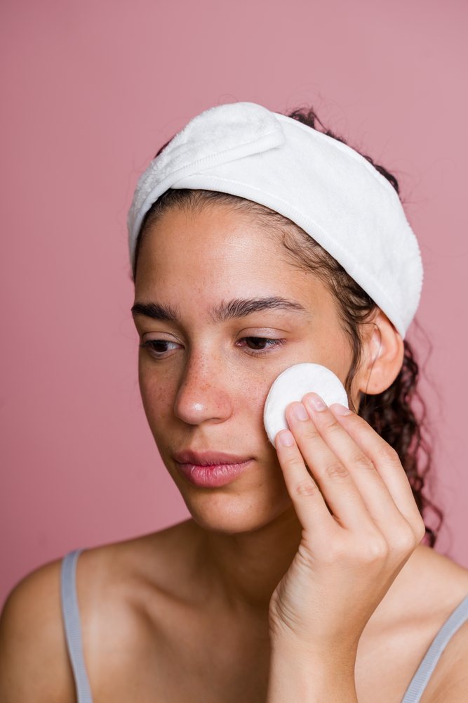 Young woman with natural skin applies skincare using a cotton pad while wearing a white headband against a pink background, beauty shoot, Spain.