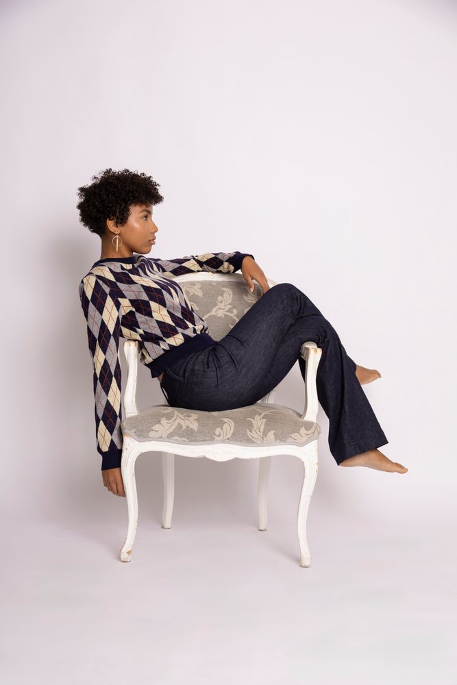 Woman wearing an argyle sweater and jeans sits barefoot on an upholstered chair in a studio with white background, fashion, young culture.