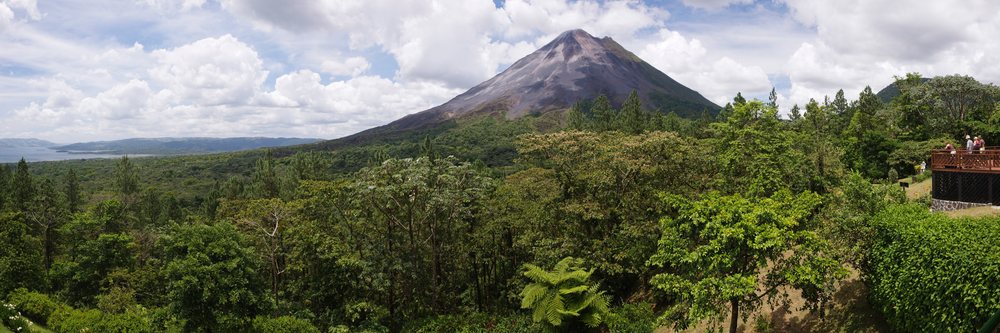 Scenic view of Arenal Volcano and volcanic cone mountain framed by dense forest and clouds from a wooden observation deck with hikers.