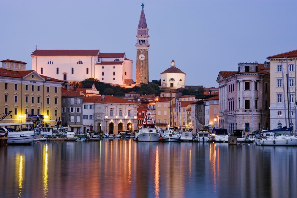 Town of Piran harbor with boats, sailboats, historic buildings, and a church tower reflected in calm water, Dusk, Slovenia.