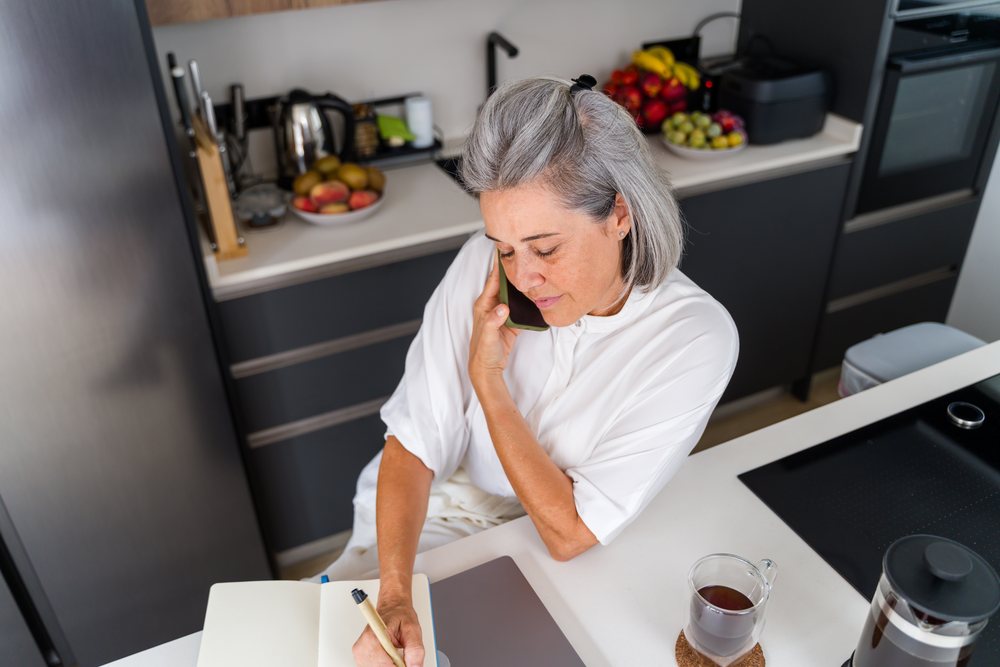 Mature woman with gray hair talking on phone and writing in notebook in modern kitchen, Spain.