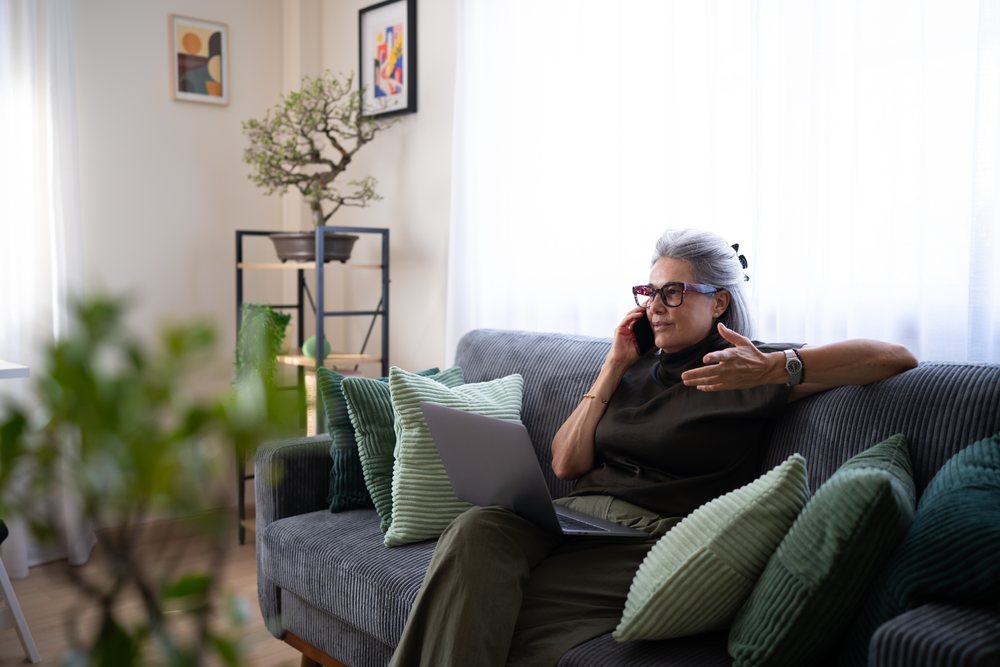 Mature woman with gray hair talks on the phone while using a laptop seated on a sofa indoors, Spain.