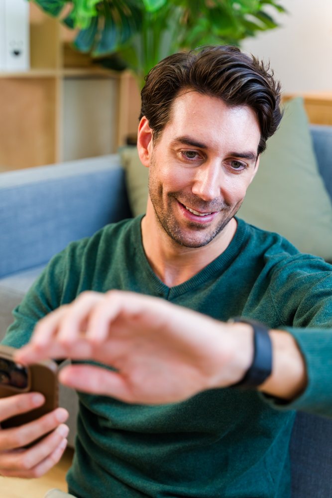 Man wearing a green sweater sits on a couch smiling while checking his smartwatch in a living room, Spain.