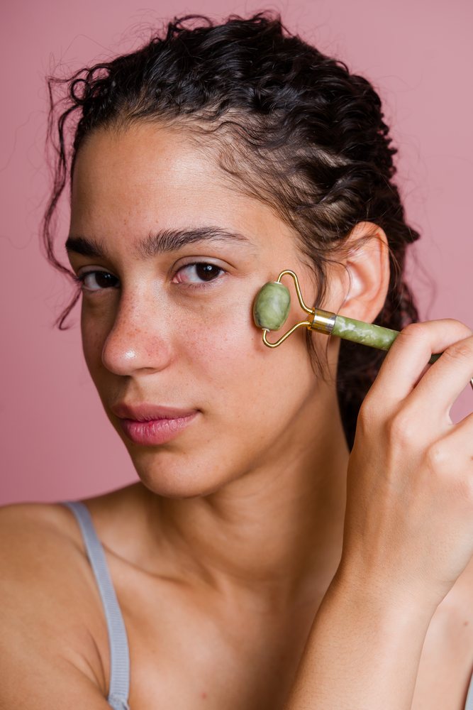 Woman performing facial massage using a jade roller against a pink background during a beauty shoot, Spain.