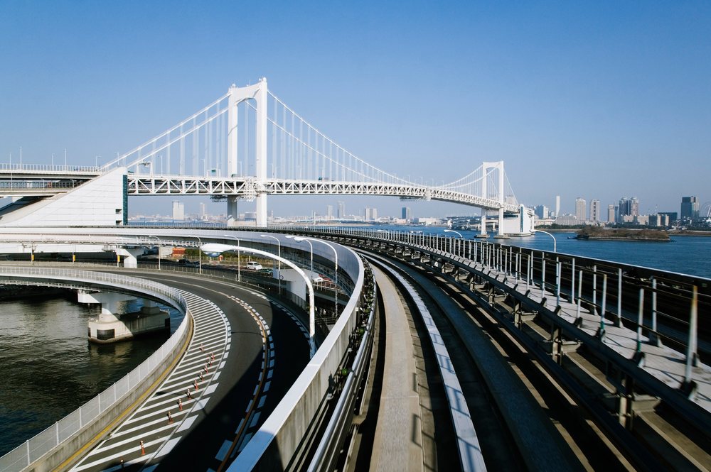 Rainbow Bridge and curved Yurikamome Monorail tracks span water with Tokyo skyline in background under blue sky, Japan.