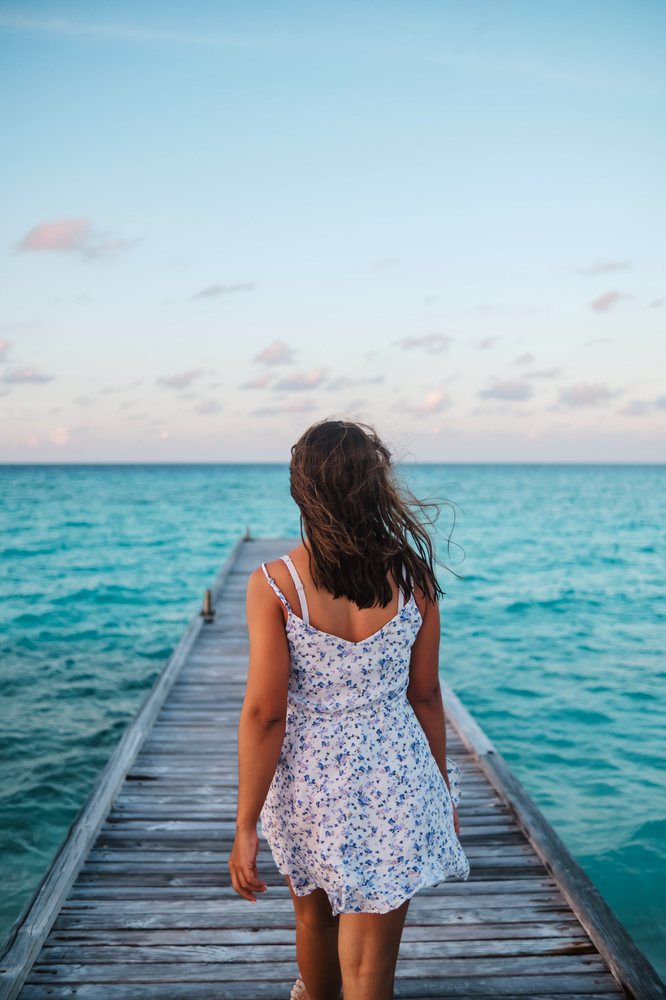 A woman wearing a blue floral summer dress walks on a wooden pier toward the turquoise ocean in the, Maldives.
