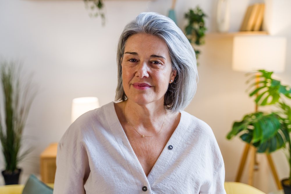 Mature woman with gray hair smiling indoors in a modern living room with houseplants and a lamp, Spain.