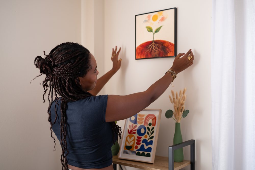 A woman is hanging a framed painting of a sprouting plant on the wall in a cozy home office decorated with contemporary art and dried plants, remote work, student, Spain.