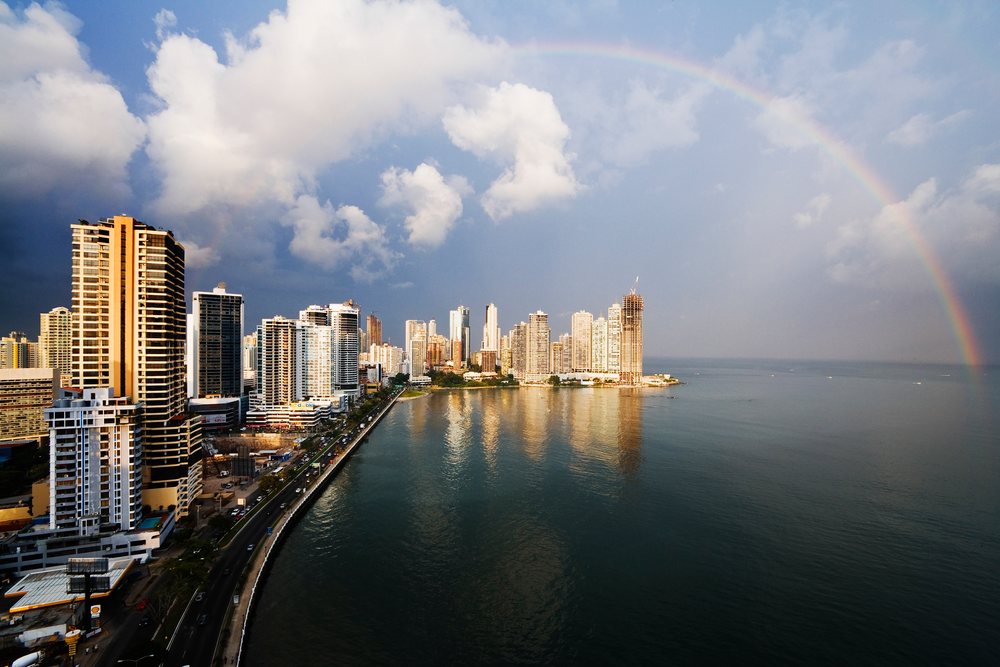 Panama City skyline with high-rise buildings and a rainbow arching over the ocean along the urban waterfront.