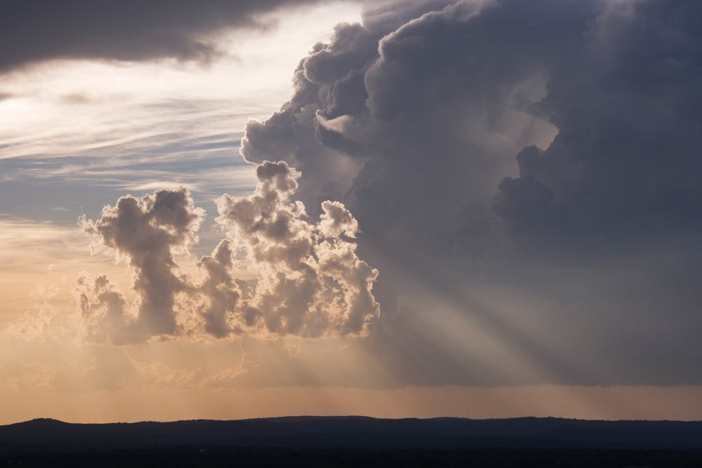 Storm clouds with sun rays visible above Texas hills in the evening sky.