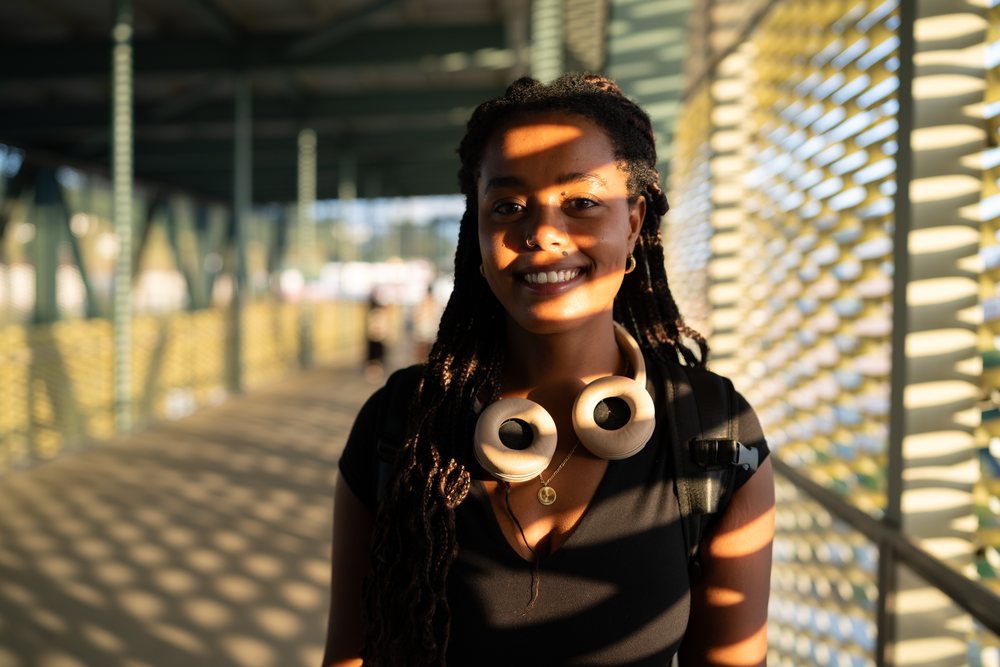 A young woman smiling with headphones around her neck on a sunlit modern pedestrian walkway, urban, Santiago De Compostela, Spain.