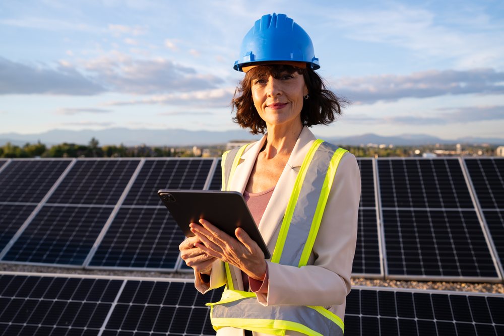 Smiling woman wearing blue hard hat and safety vest holds a black tablet outdoors with solar panels and mountains behind her, Madrid, Spain.