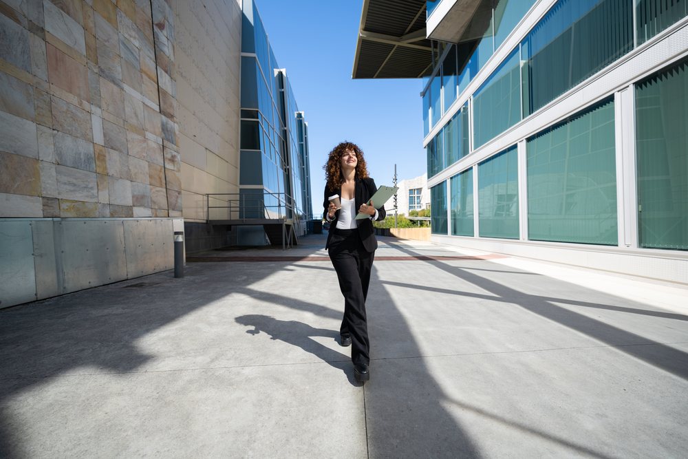 Woman walking outdoors with clipboard and coffee cup between modern buildings, businesswoman, traveling executive, Spain.