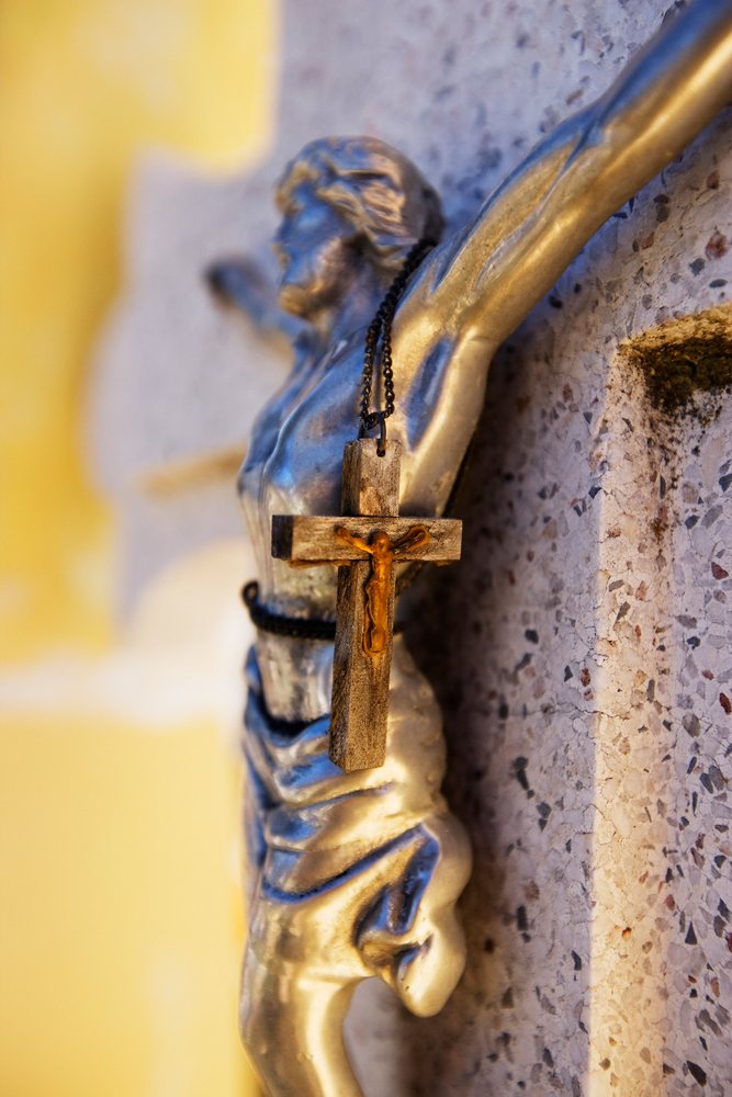 Close-up showing a metal crucifix statue with a wooden cross pendant hanging on it, affixed to a stone headstone.