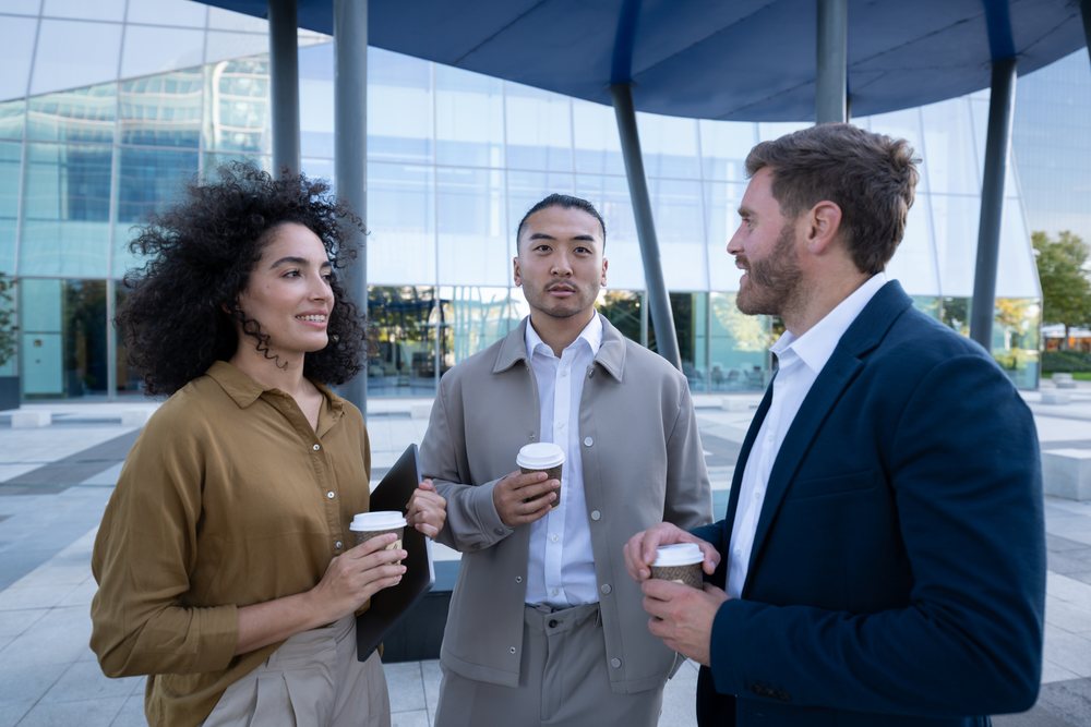 Three young professionals holding disposable coffee cups and talking outside a modern glass office building in an urban setting, Madrid, Spain.