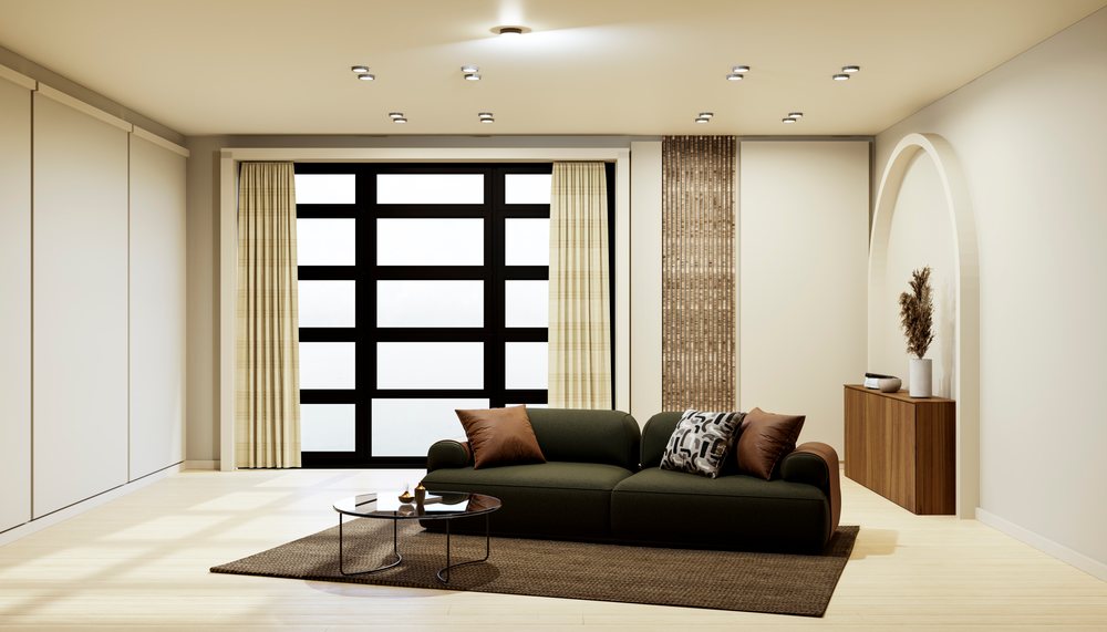 Modern minimalist living room featuring a green sofa with pillows, coffee table on a brown rug, wooden cabinet with vase and dried plants, and natural light from a large window.
