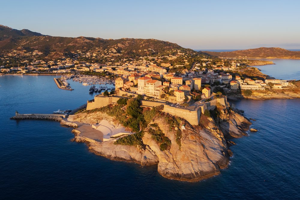 Citadel of Calvi on rocky coastline overlooking the Mediterranean Sea at Sunrise with harbor and mountains in the background, Travel Photography, Corsica, France.