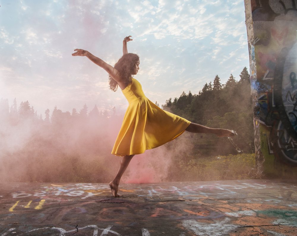 Female dancer in a yellow dress performs barefoot outdoors near graffiti walls with fog and trees visible in the background, USA.