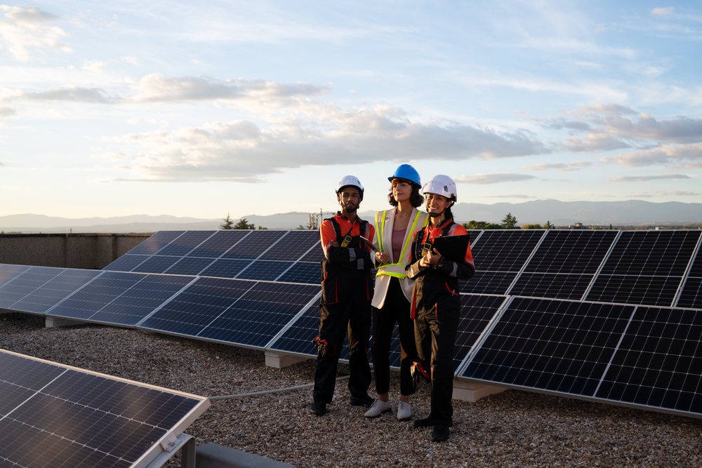 Three engineers wearing helmets and protective gear inspect solar panels outdoors with mountains and blue sky in the background, Madrid, Spain.