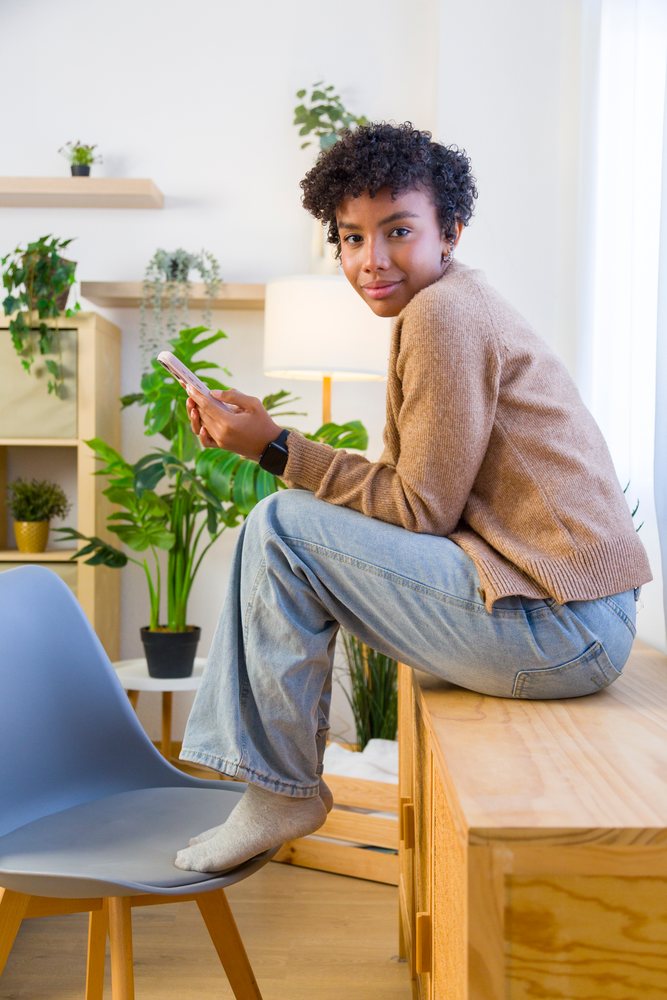 Young woman sitting on a wooden cabinet using a smartphone in a modern home interior with plants and a lamp.