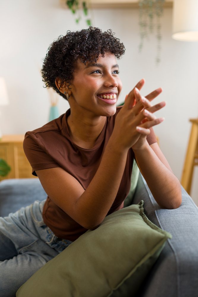 Young woman with curly hair sits smiling on a gray sofa with a green pillow in a home interior.