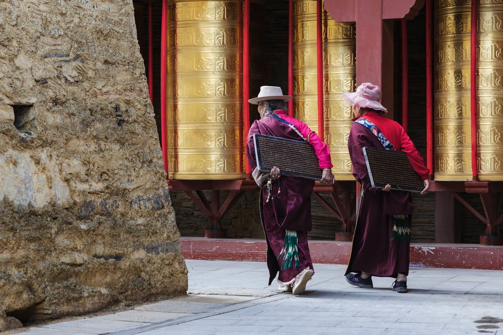 Two Tibetan women carry inscribed wooden tablets near golden prayer wheels at a temple, China, Tagong, Sichuan Province, People's Republic of China.