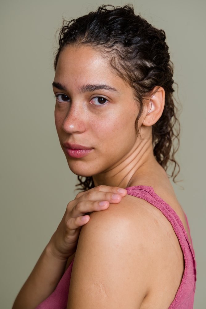 Young adult woman with curly hair and freckles touches her shoulder, looking at the camera in a studio, beauty shoot, Spain.