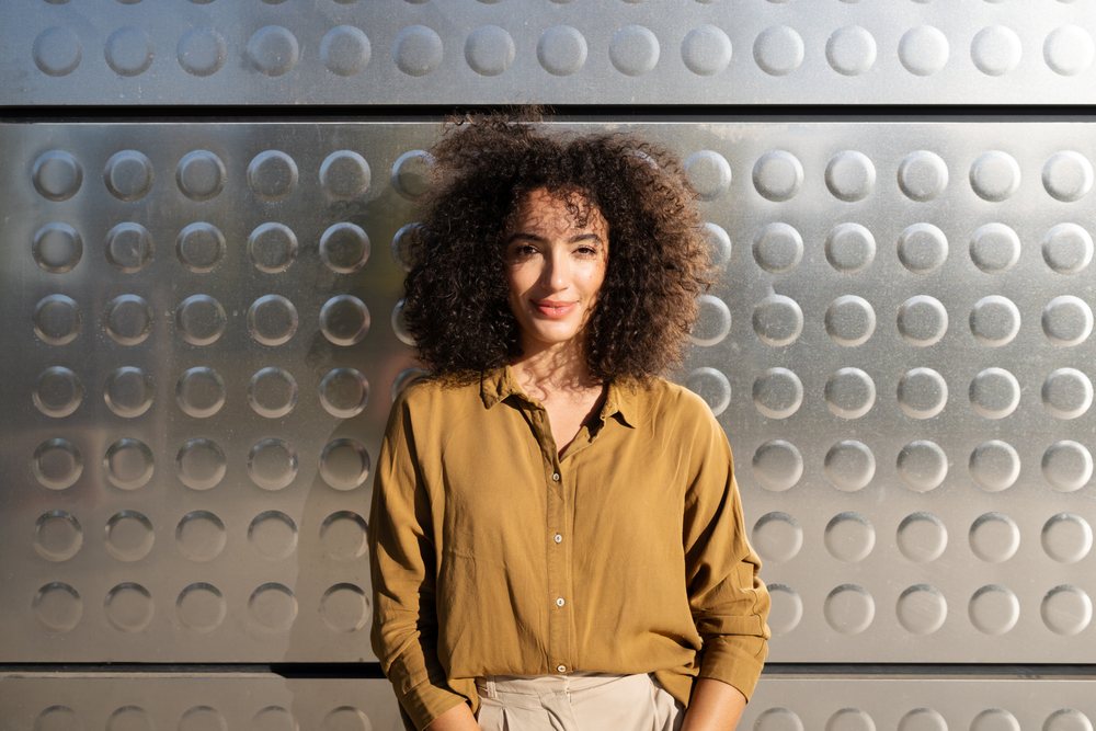 Woman with curly hair wearing a tan shirt stands outdoors against a metal wall with circular texture in sunlight, Madrid, Spain.