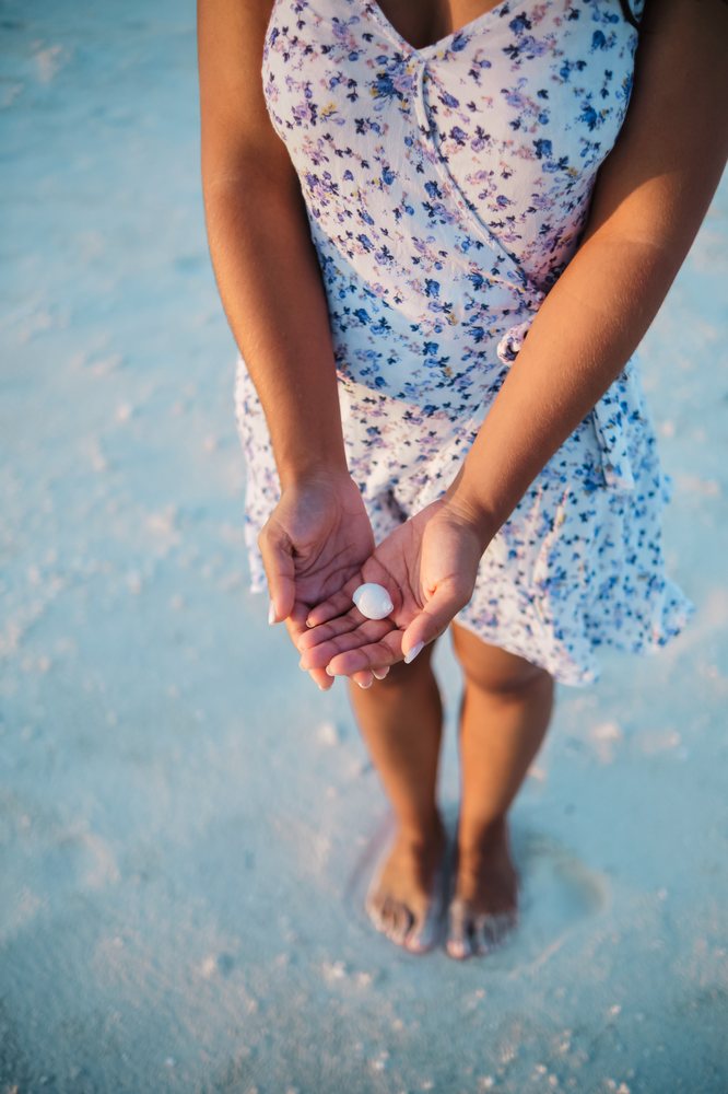 A woman in a floral summer dress stands barefoot on the sand holding a small white shell in her hands on a beach in the, Maldives.