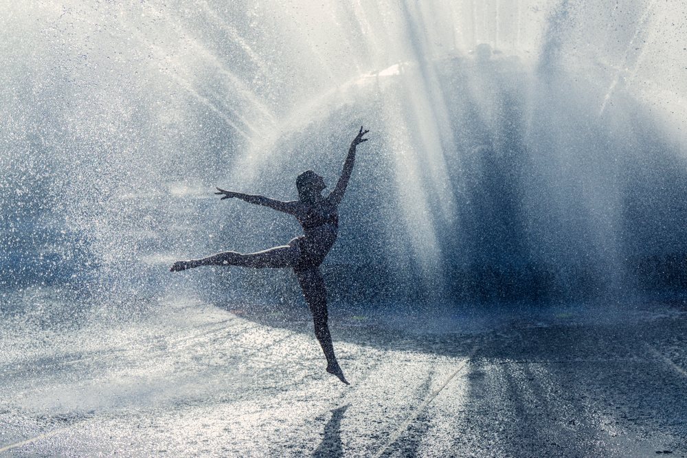 female dancer performing a ballet pose in water spray creating a dynamic silhouette and water droplets in an outdoor stage setting, partner, acrobatics, yoga, USA.