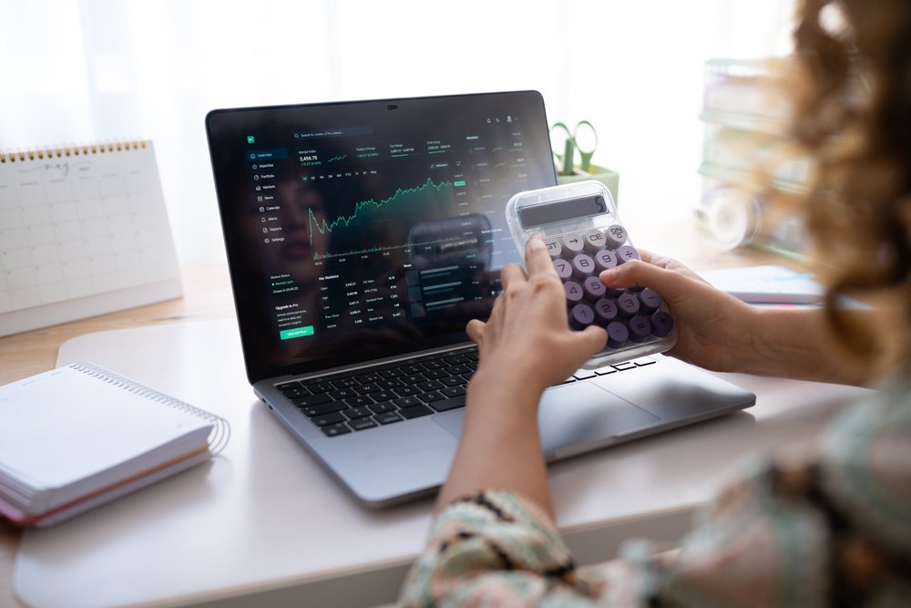 Woman holding calculator while viewing stock market graph on laptop at a desk in Spain workspace, home, girl.