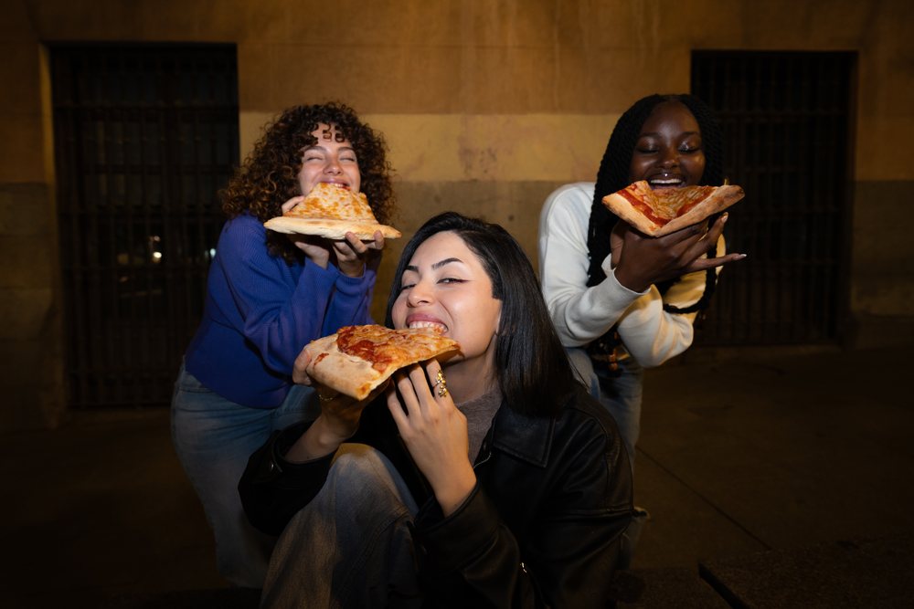 Three friends smiling and eating cheese and pepperoni pizza slices outdoors at night, Madrid, Spain.