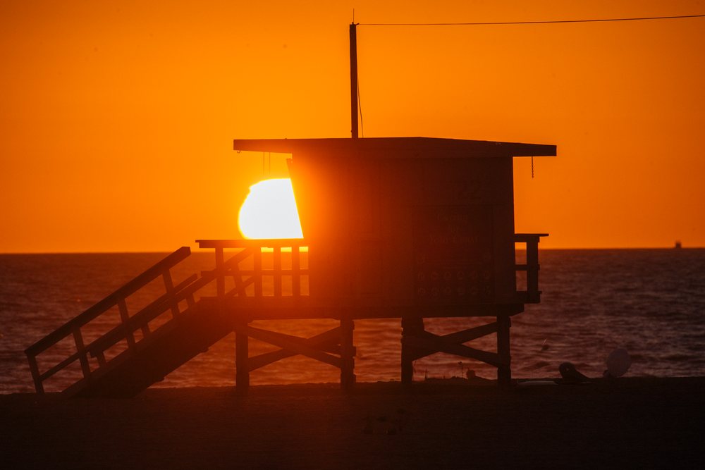 Sunset glows behind a silhouetted lifeguard tower on the Santa Monica beach shore at evening, CA, USA.