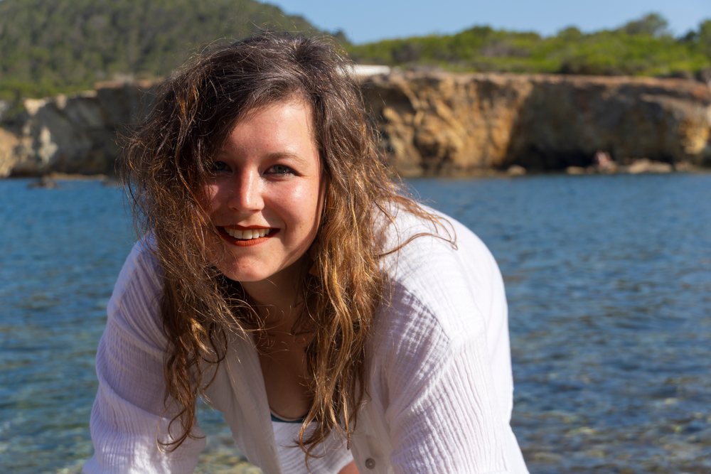 Woman smiling outdoors near a rocky shore beside blue water, photo shoot, Ibiza, Spain.