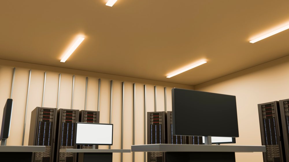 Computer monitors on desks in front of server racks in a data center room with beige walls and shelving.