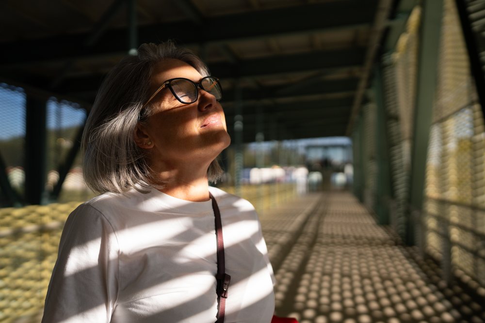 Woman with gray hair and eyeglasses relaxing in sunlight on a metal bridge pathway outdoors, Spain.