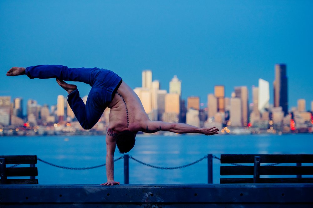 Shirtless man with tattoos performing a one-hand handstand yoga pose on the Seattle waterfront at dusk with city skyline in background, partner acrobatics, USA.