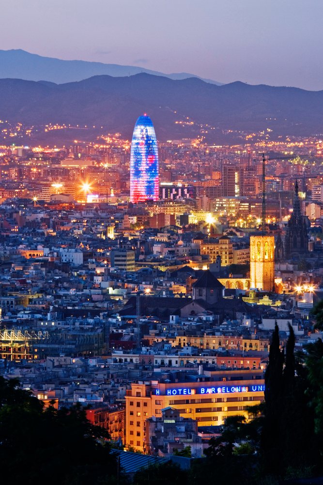 Dusk cityscape of Barcelona with illuminated buildings, mountains in the background, and vibrant urban lights.