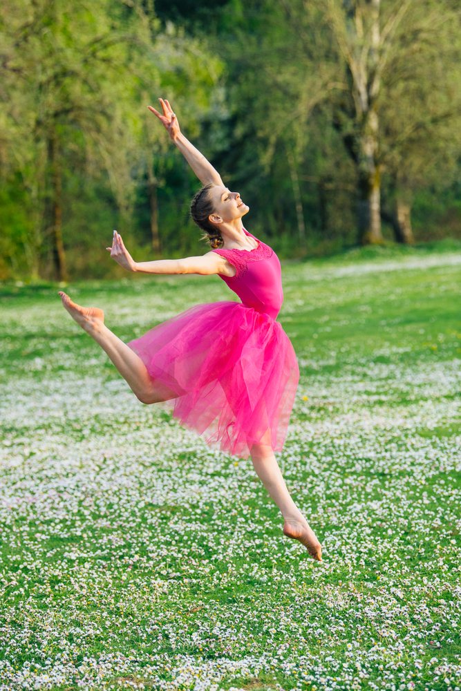 Female ballet dancer in a pink tutu leaps gracefully in a flower-covered green outdoor field in spring with trees in the background, USA.