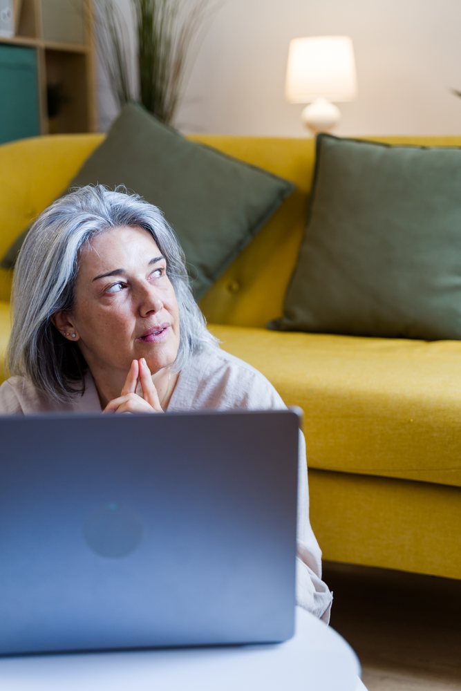 Gray-haired woman is thinking while using a laptop in a living room with a yellow sofa and lamp, Spain.