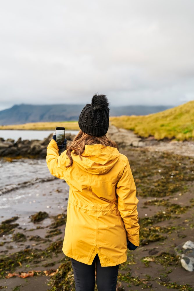 A white woman takes a smartphone photo on a rocky shoreline in a beach