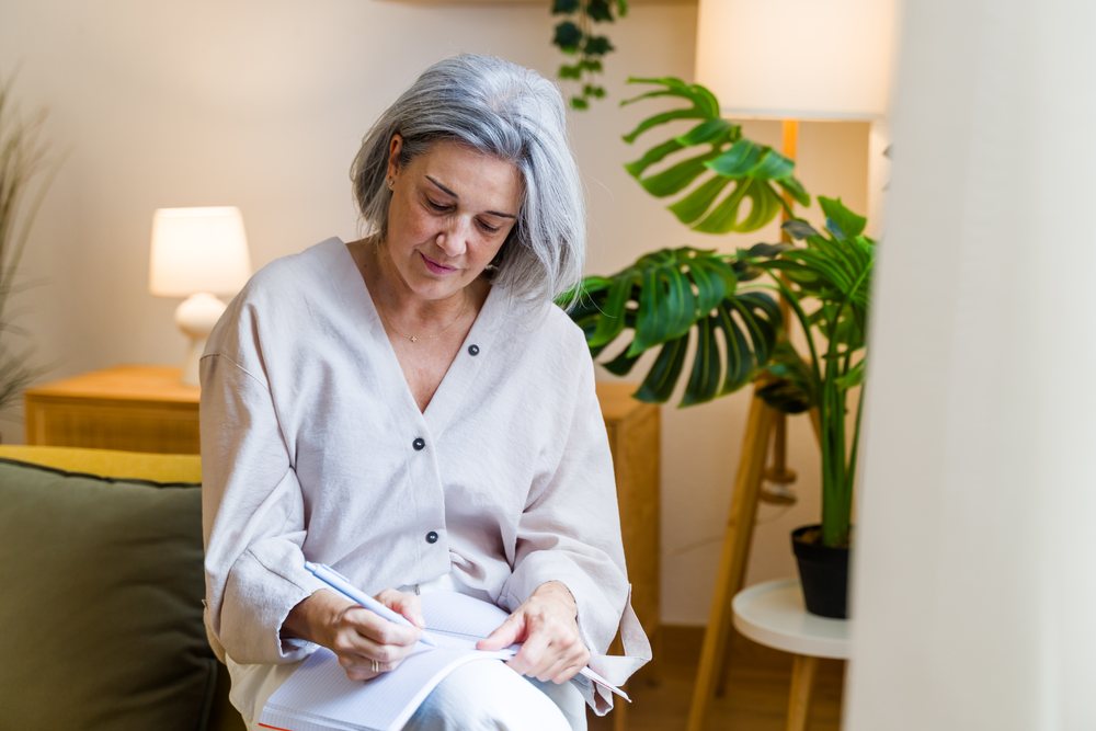 Woman with gray hair writes in a notebook indoors beside a lamp and plant in a cozy home, Spain.
