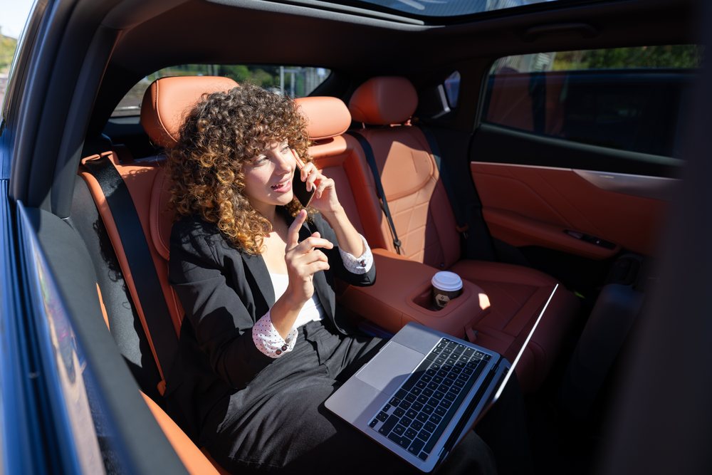 Businesswoman in black suit working on a laptop and talking on phone inside a car with orange leather seats and a coffee cup in daylight, remote work, Spain.
