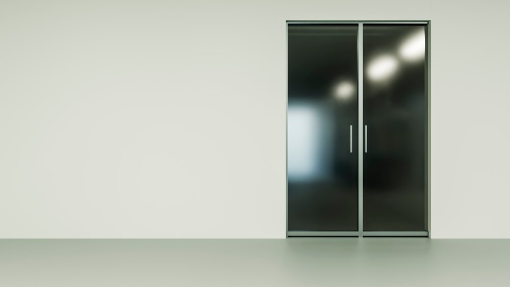 Closed sliding glass door with metal frame and vertical handles in a minimalist empty room with white walls and smooth floor, showing reflections on the glass surface.
