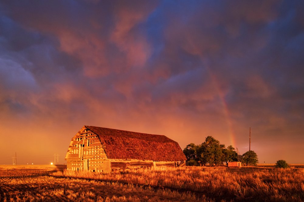 Old barn stands in a rural field at sunset with a rainbow arching above and storm clouds in the sky, Storm Chasing, Weather, Environment, Eva, Oklahoma, USA.