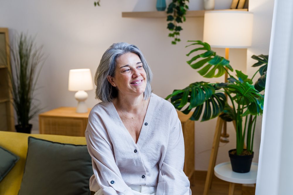 Middle aged woman with gray hair smiling while seated on sofa in a modern living room with plants and lamp, Spain.